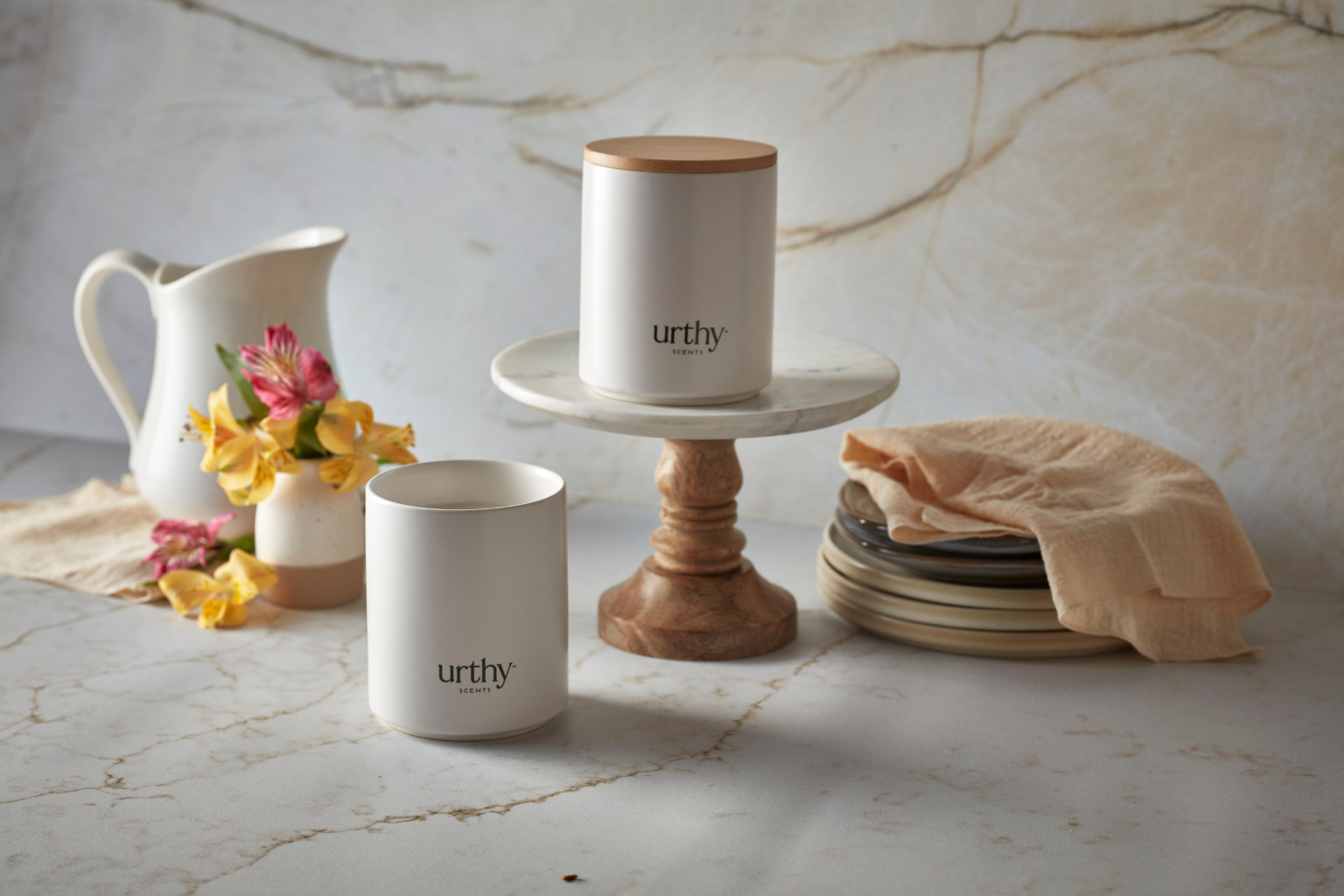 Two white Urthy candle jars with wooden lids are styled on a marble surface with a white pitcher, yellow flowers, stacked plates, and a beige cloth. One jar sits on a marble-and-wood pedestal and the other is placed beside it.